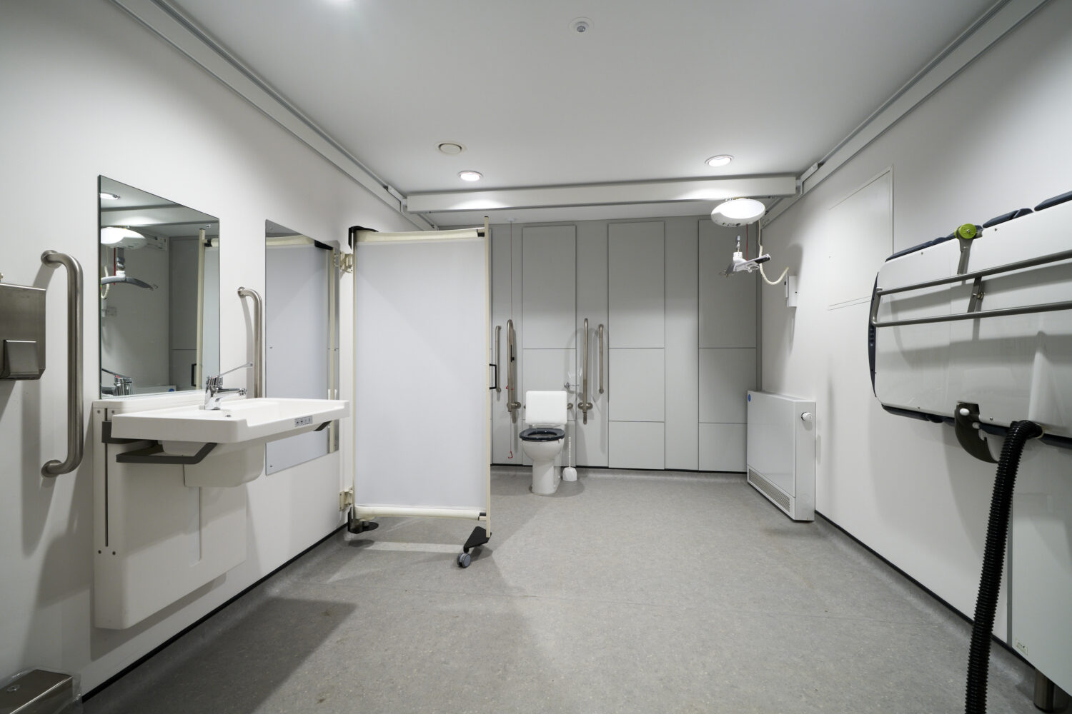 Modern, accessible restroom with sleek white fixtures, grab bars, and ample space. Includes a fold-down changing table and a privacy screen.