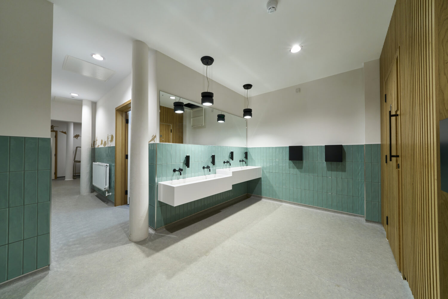 Modern restroom with green tiles, a long white sink, and black faucets under hanging black lights. The space feels clean and minimalist.