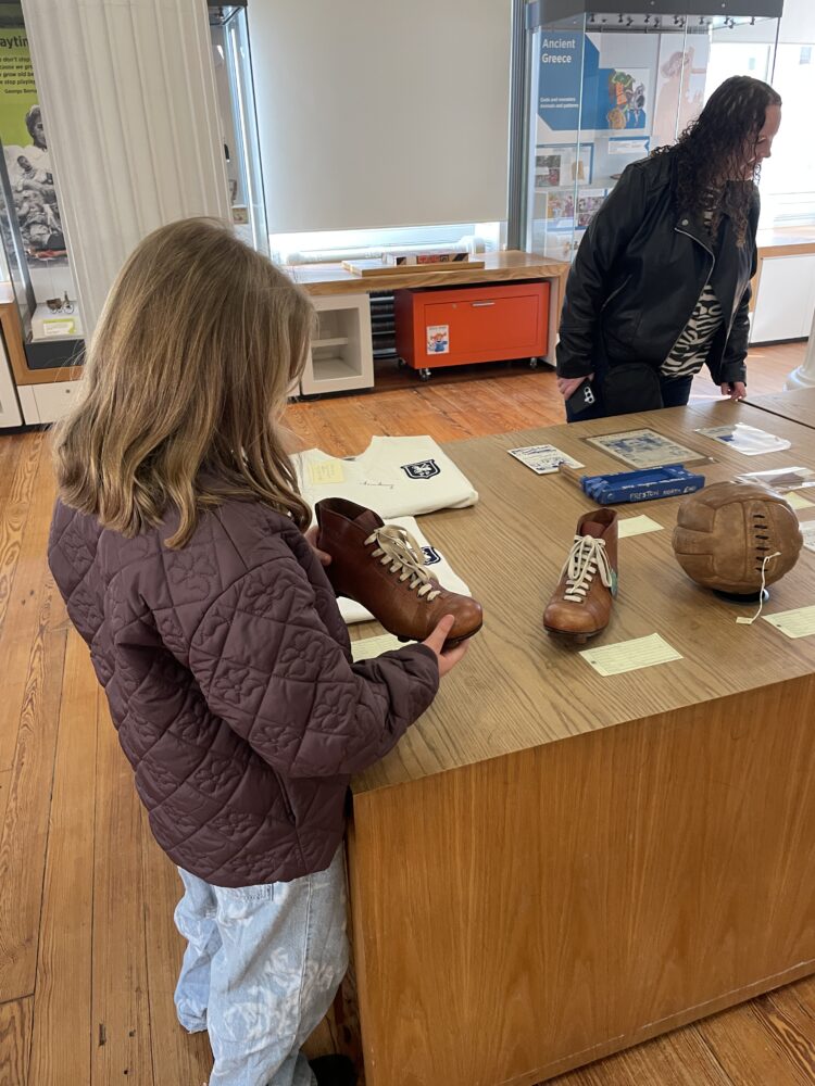 A child in a purple jacket examines vintage soccer cleats displayed on a wooden table in a museum, evoking curiosity and a sense of history.