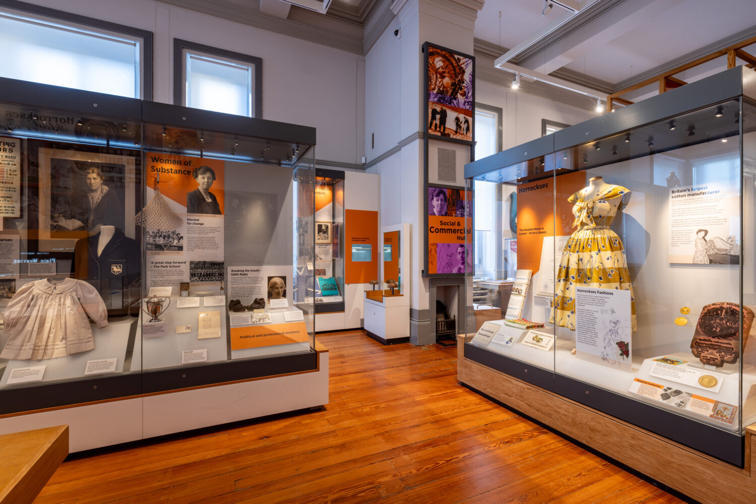 Museum exhibit room featuring historical women's clothing displays. Bright orange and informative panels highlight women's contributions, creating an educational and inspiring atmosphere.
