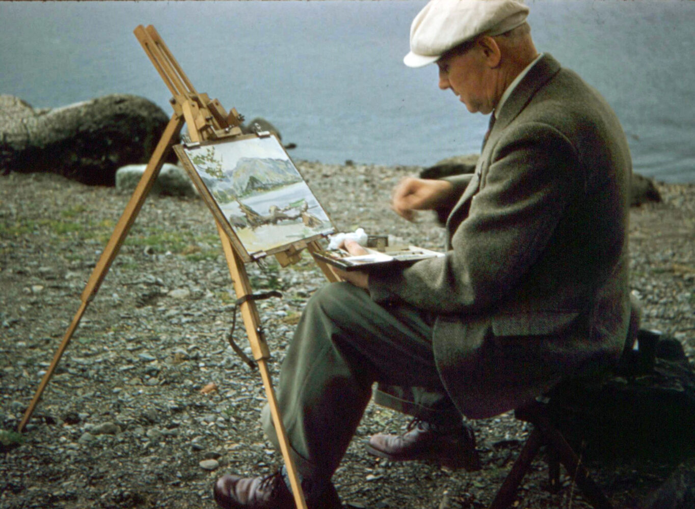 An image of James Brindle sat near a lake on a stone shore, painting the landscape.