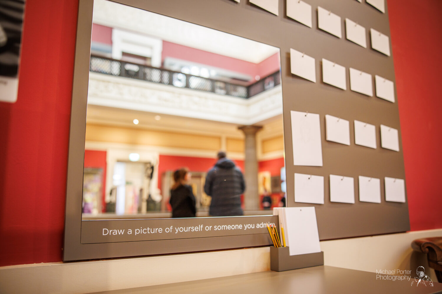 A museum wall with blank papers and pencils invites visitors to draw portraits. Reflected in a mirror are two people in an ornate gallery. The mood is inviting.