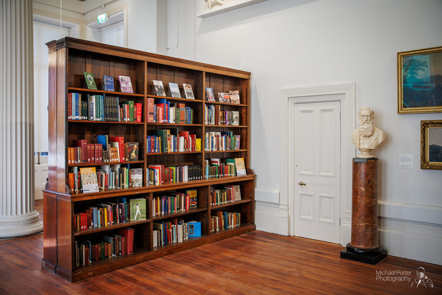 A tall wooden bookshelf filled with colorful books stands in a bright room. Nearby, a white sculpture bust is placed on a pedestal next to a closed door.
