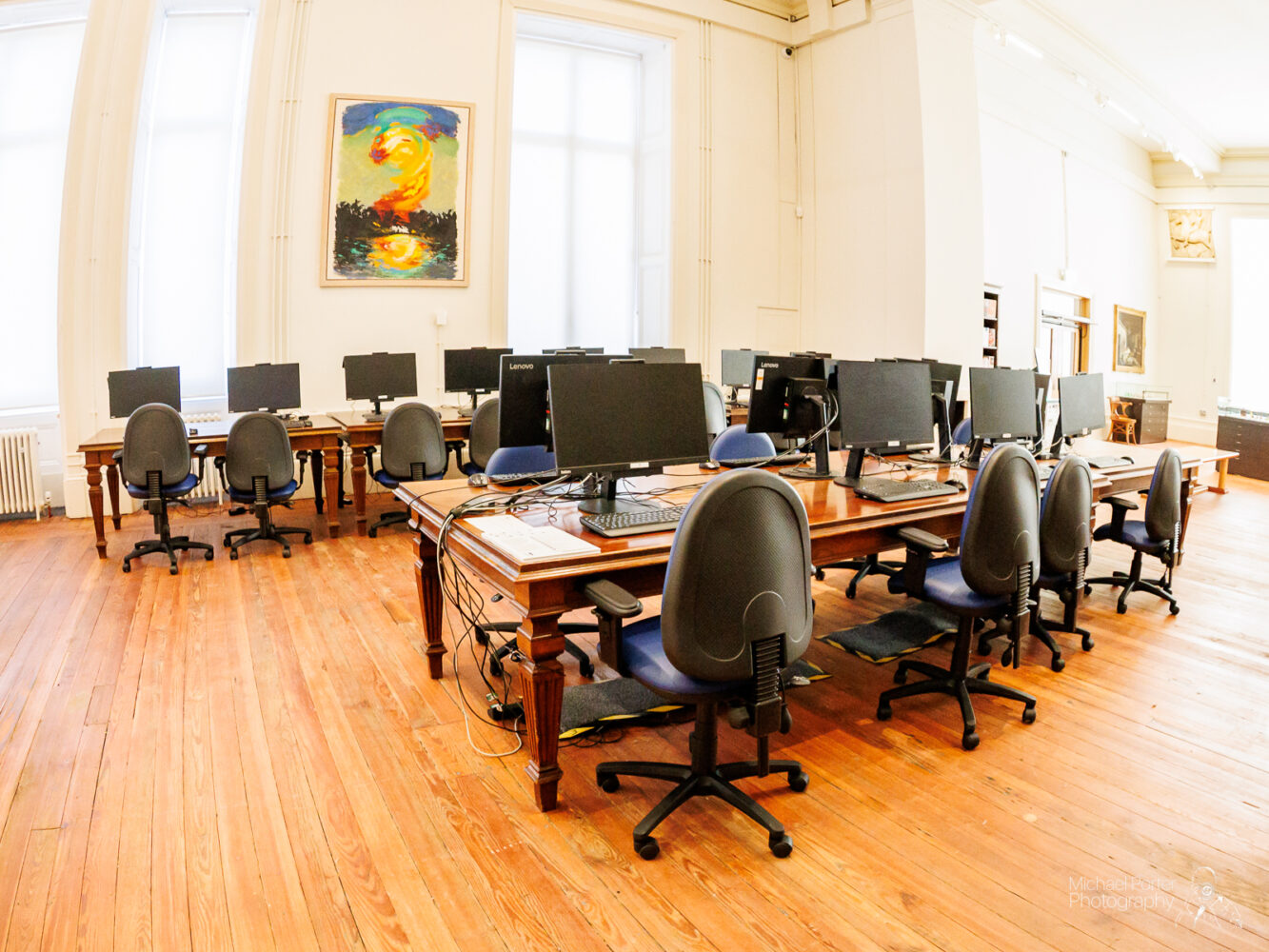 Spacious computer lab with wooden floors and large tables, lined with monitors and swivel chairs. A colorful abstract painting hangs on the wall, creating a modern, artistic vibe.