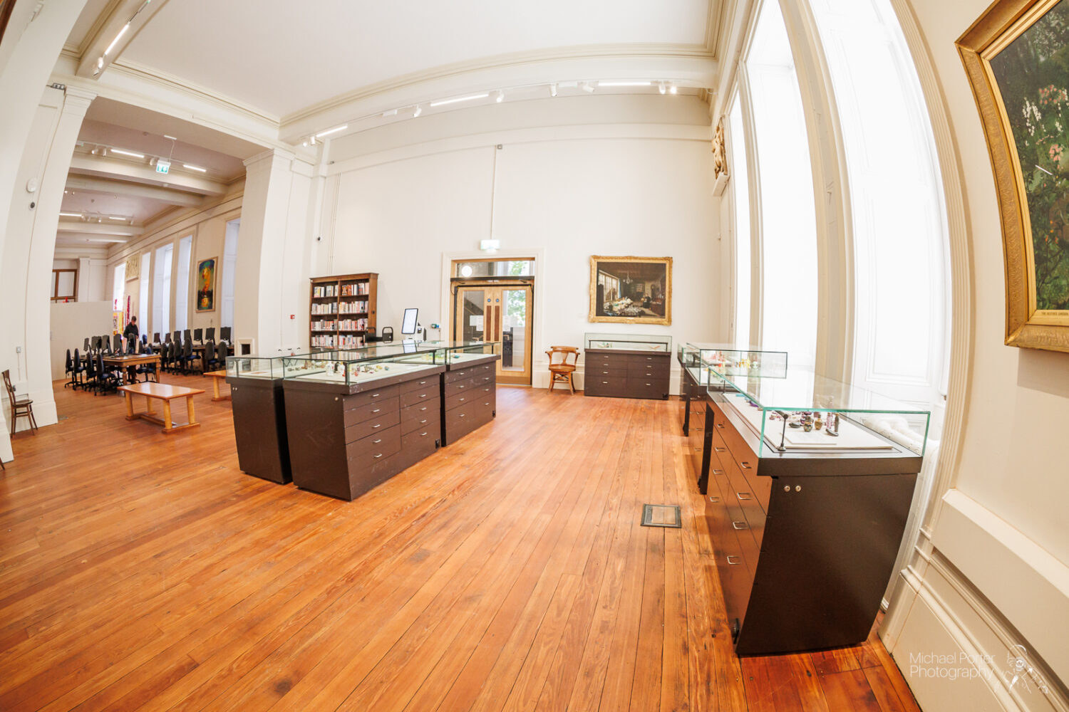 Spacious art gallery with wooden floors, large windows, display cases, and framed paintings. Bright, elegant, and inviting atmosphere.