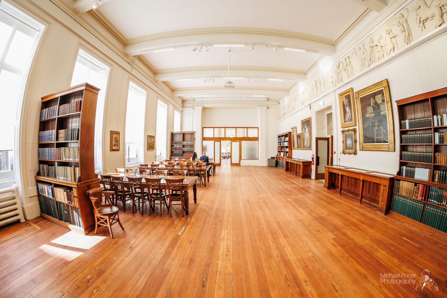 Spacious library room with tall windows, wooden floors, tables, and chairs. Shelves of books and framed paintings adorn the walls, creating a serene atmosphere.