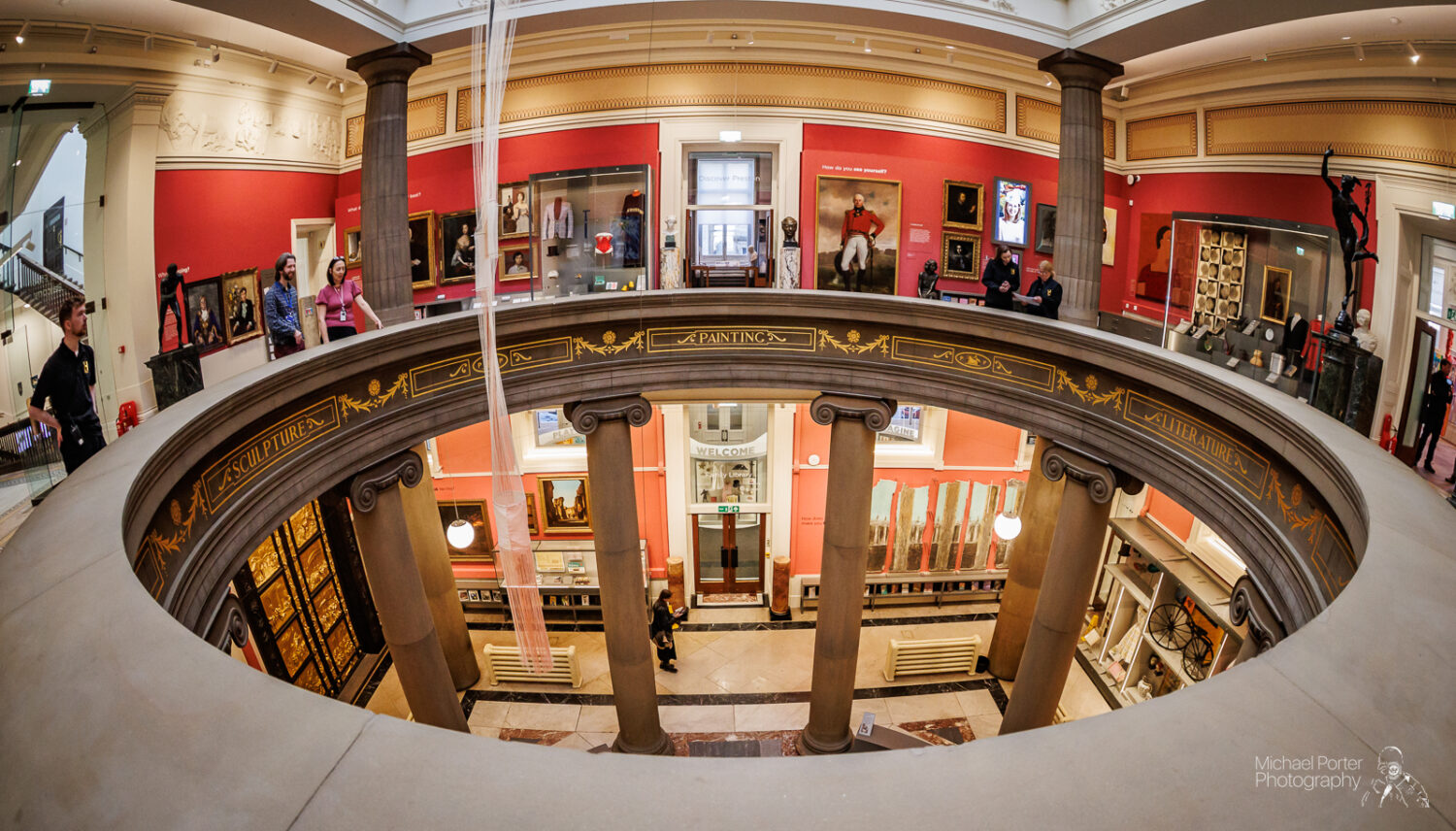 A fisheye view of an ornate art gallery with red walls, showcasing paintings and sculptures. Visitors explore the gallery, creating a lively, cultural atmosphere.