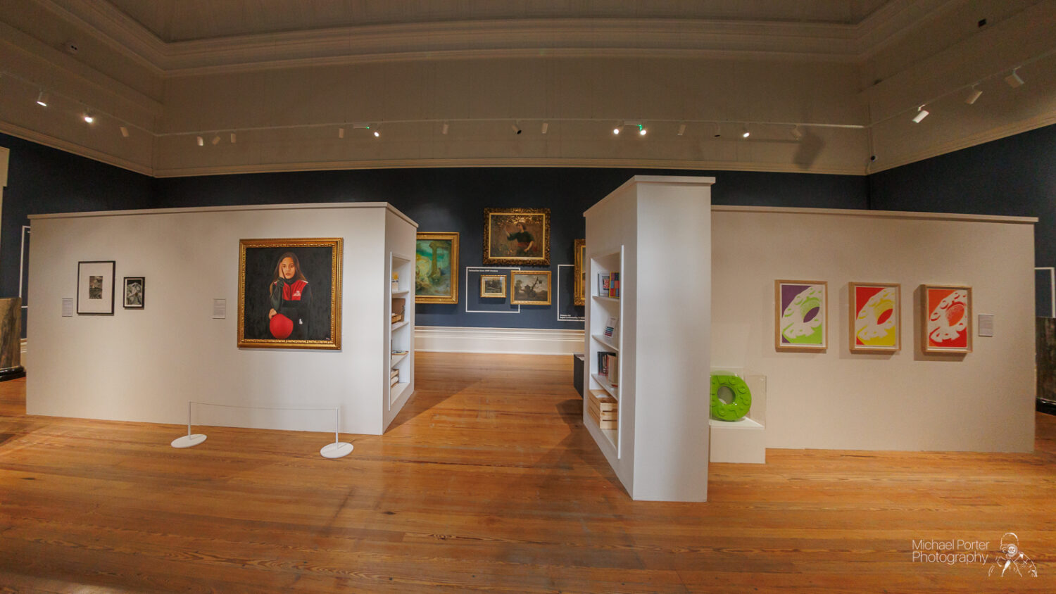 A spacious art gallery room with wooden floors and blue walls displays various artworks. Two white partitions showcase colorful framed paintings and photographs.