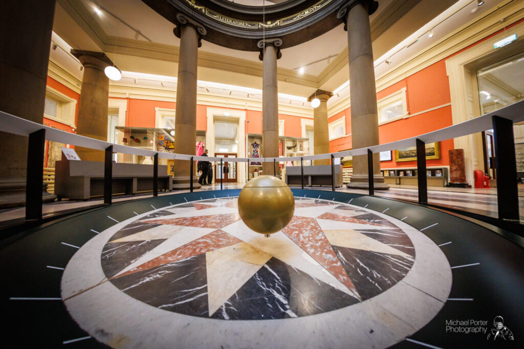 A golden pendulum swings above a circular, marble compass design on the floor of a grand, columned hall with red walls and arched doorways.