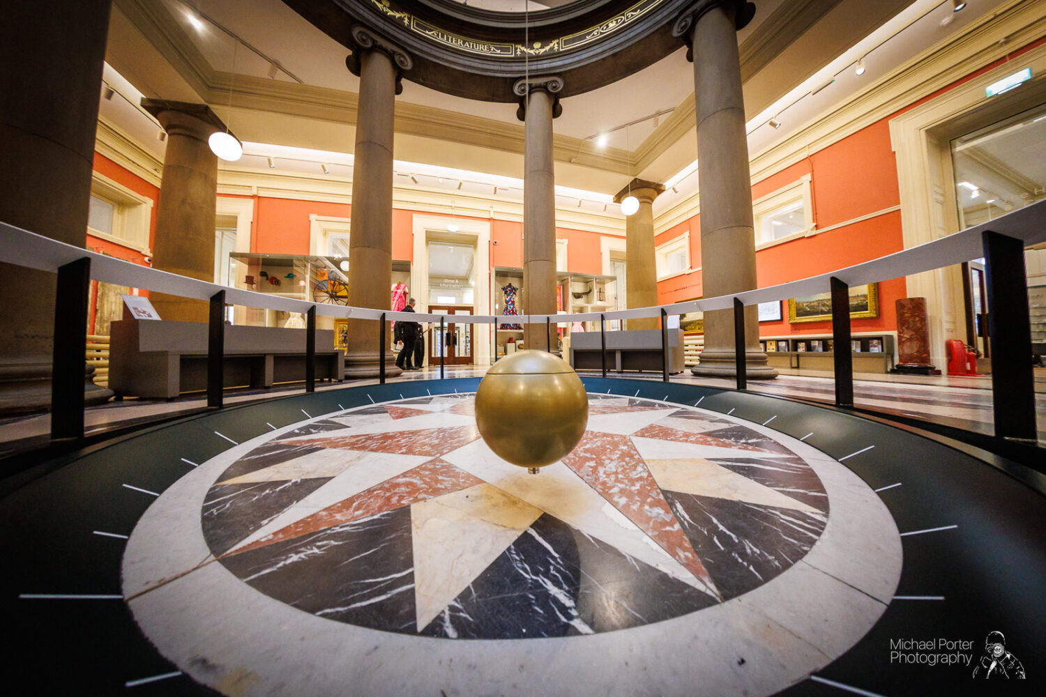 A golden pendulum swings above a circular, marble compass design on the floor of a grand, columned hall with red walls and arched doorways.