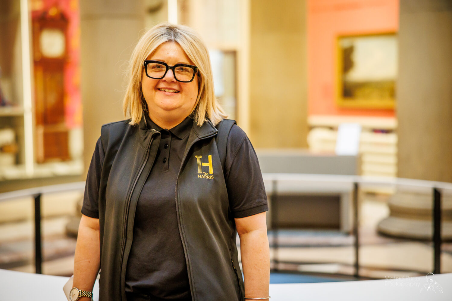 A smiling person with blonde hair wearing glasses and a black vest stands in a warmly lit museum. Artworks and a tall clock are visible in the background.