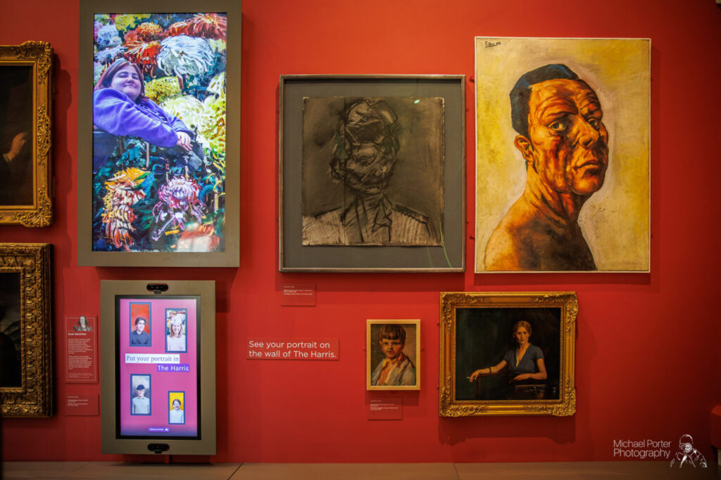 A vibrant gallery wall features digital and traditional portraits. On the left, a screen shows a woman in colorful attire beside an interactive display. Various framed artworks in diverse styles are arranged against the red background, creating an engaging and lively atmosphere.