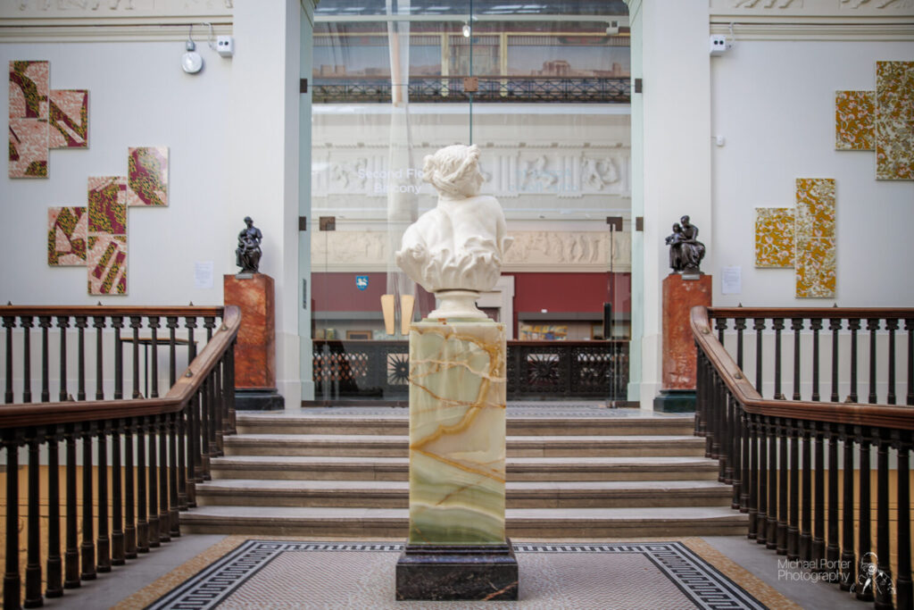 A marble bust atop a patterned pedestal stands at the center of a grand staircase in an art gallery. Sculptures and abstract wall art are displayed around.