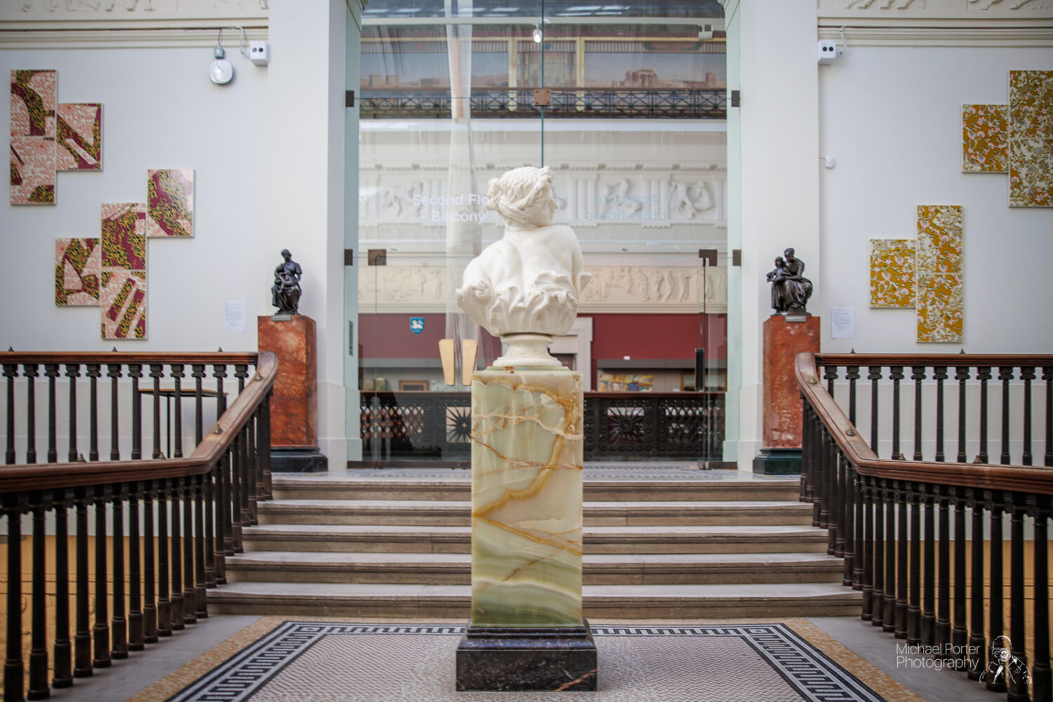 A marble bust atop a patterned pedestal stands at the center of a grand staircase in an art gallery. Sculptures and abstract wall art are displayed around.