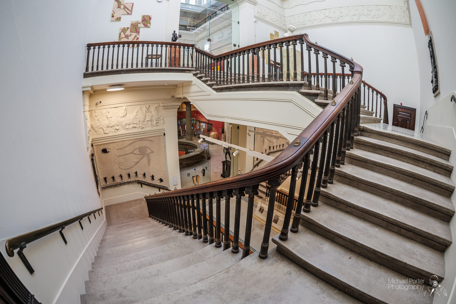 A spiral staircase with wooden railings in a well-lit museum. The walls feature Egyptian artwork, including an Eye of Horus, creating a historical ambiance.