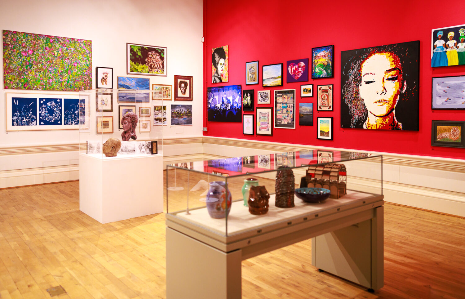 Art gallery with vibrant red and white walls showcasing diverse paintings, portraits, and multimedia art. Glass cases display ceramic pieces. Warm atmosphere.