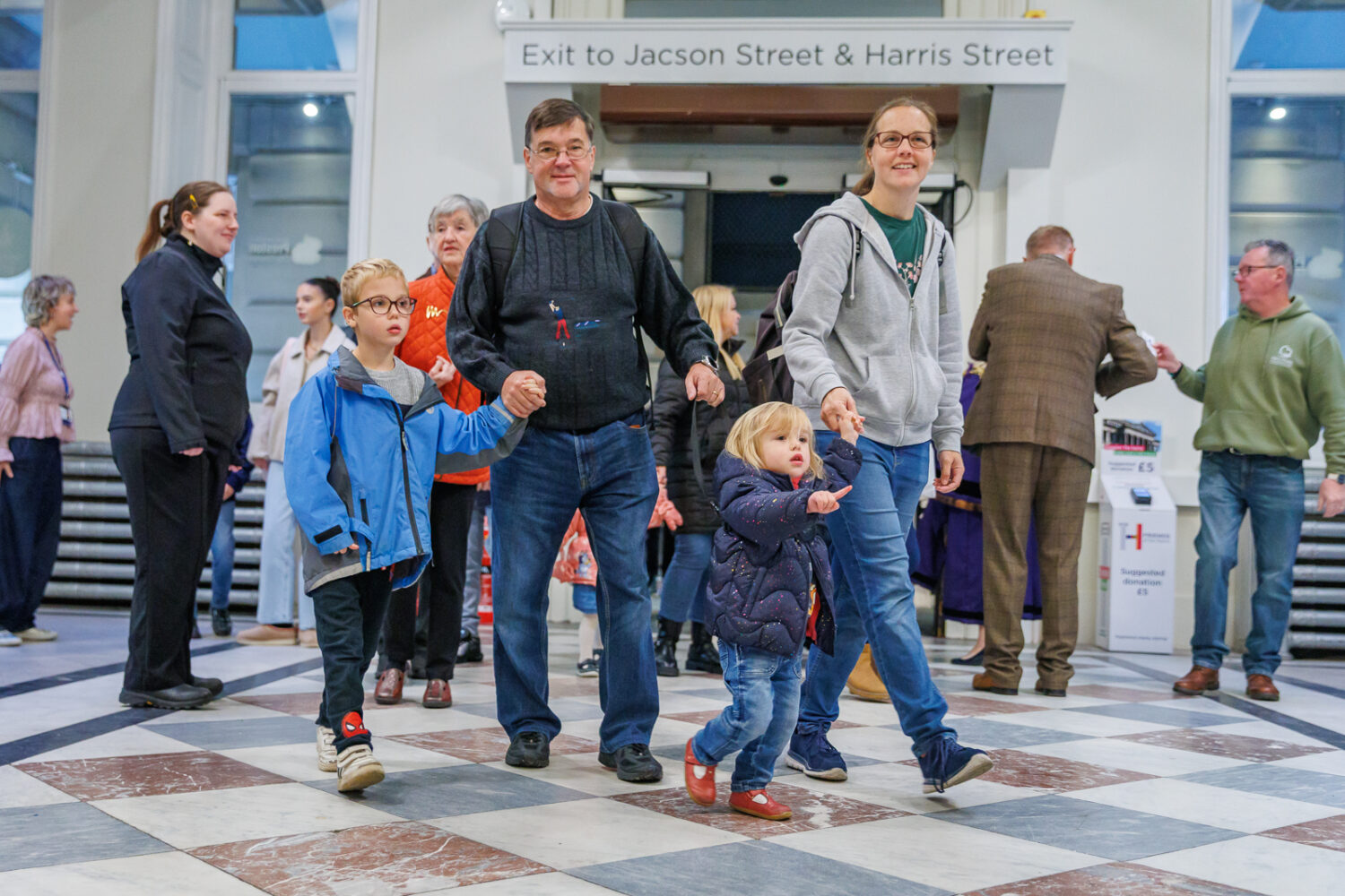A family of four, with two children, walks hand in hand through a busy station. They appear cheerful and are surrounded by other people.