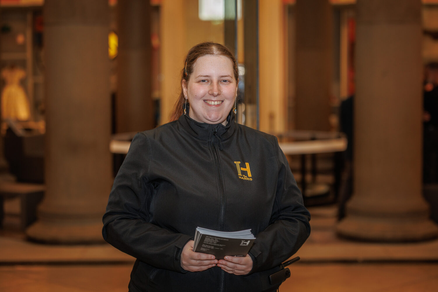 Smiling person in a dark jacket holding brochures, standing indoors. The jacket has a logo. The background features large columns and warm lighting.