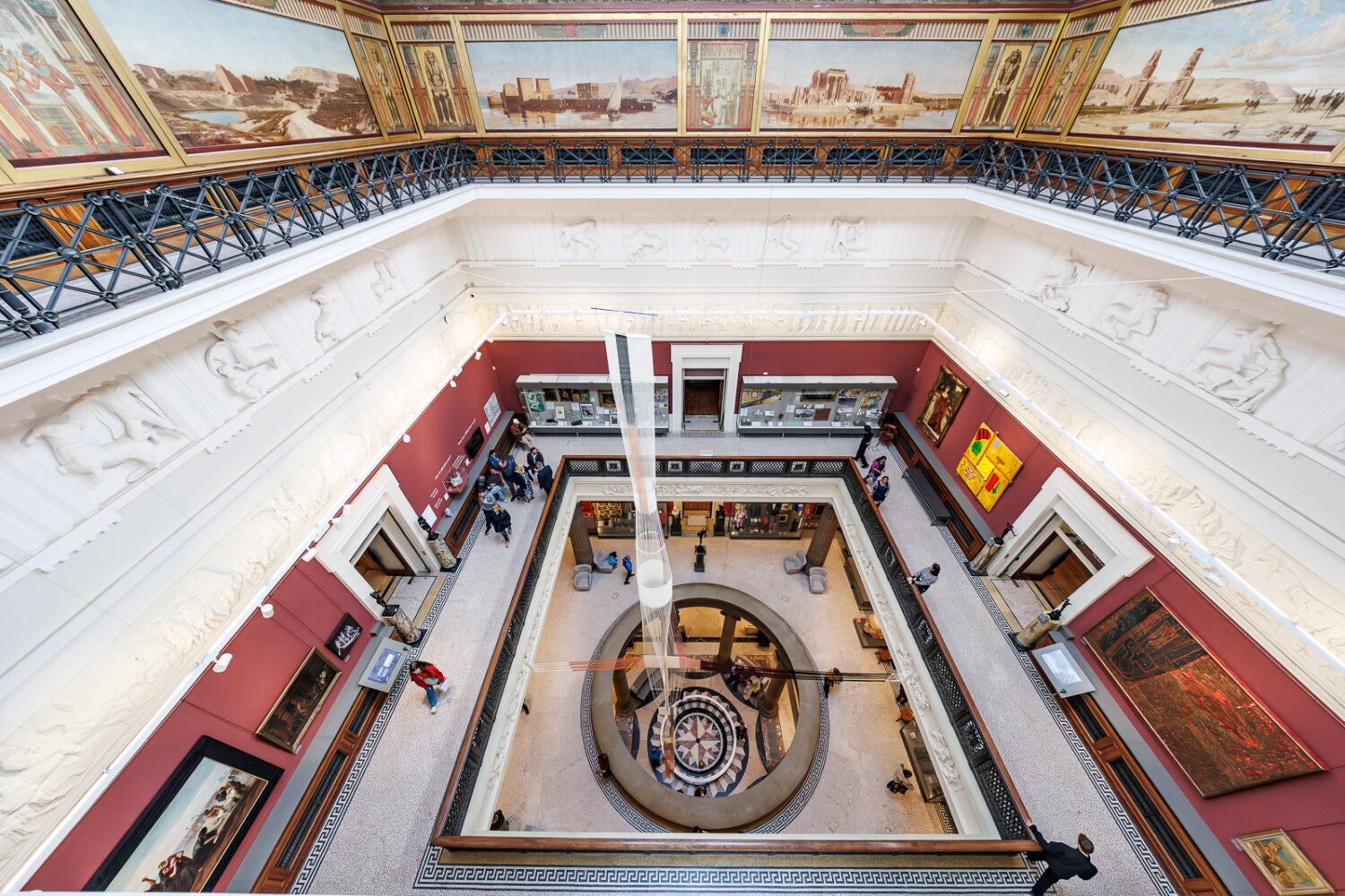 Overhead view of a grand museum atrium with ornate railings, large murals, and people admiring art on walls. Elegant, spacious, and historic ambiance.