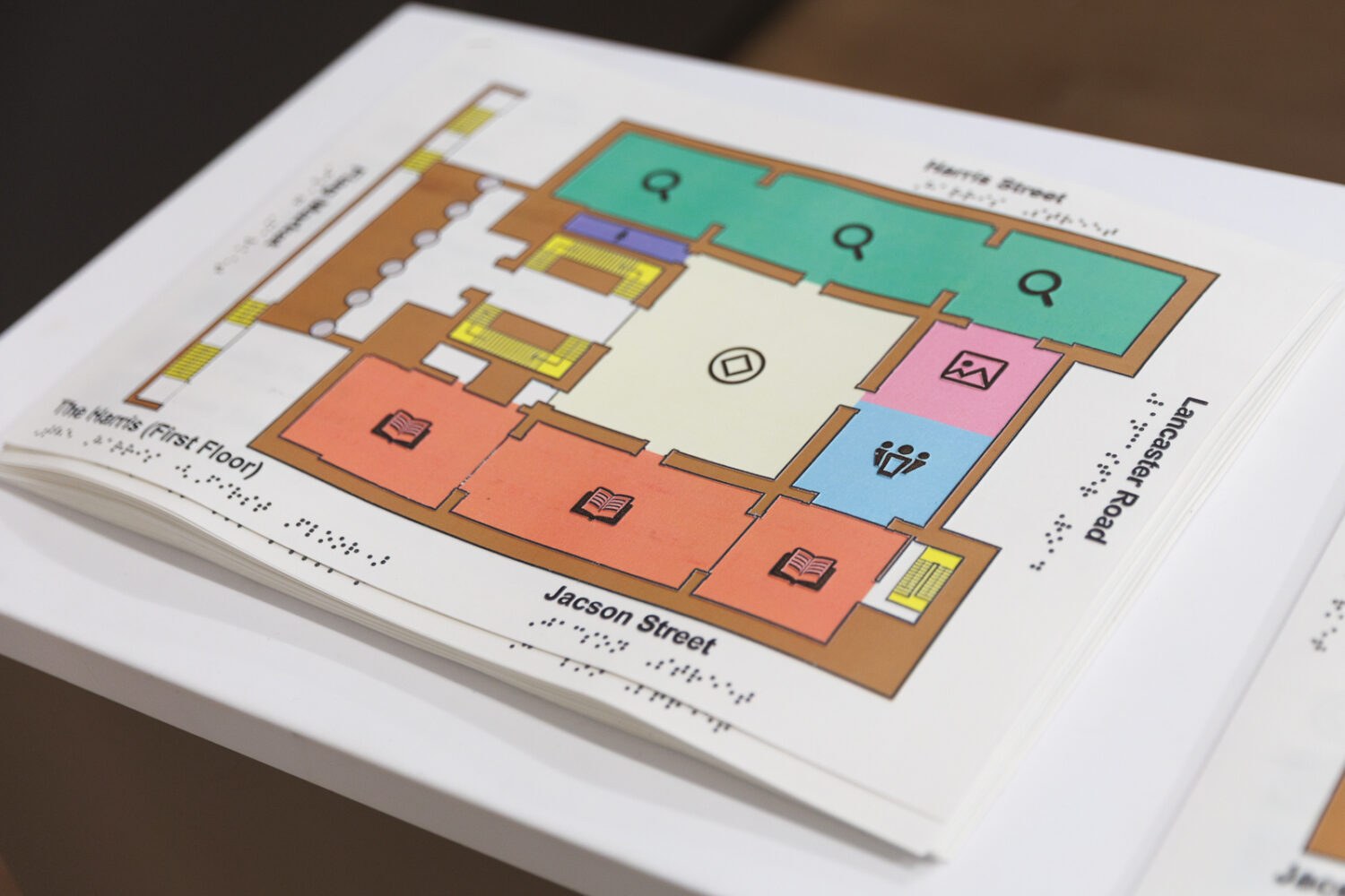 Tactile map on a table, showing a building floor plan with colored sections labeled in Braille. Bright and accessible design for visually impaired users.