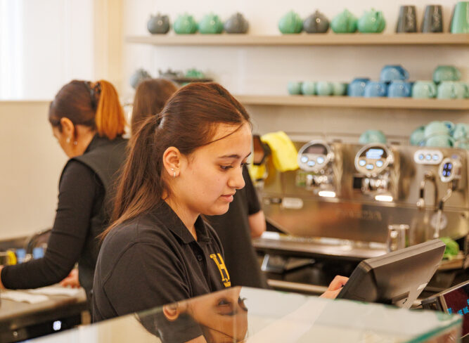 Barista at a coffee shop counter using a touchscreen. Glass display case with assorted pastries in the foreground. Shelves with teapots behind.