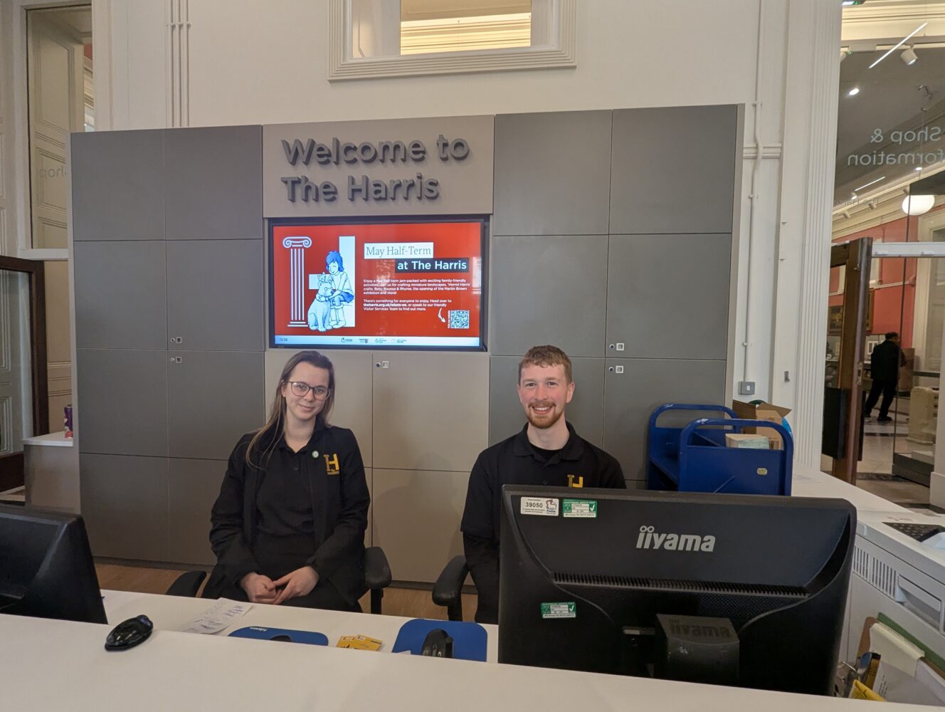 Two people behind a reception desk at The Harris. They smile warmly. A digital sign above them displays event details. Bright, welcoming space.