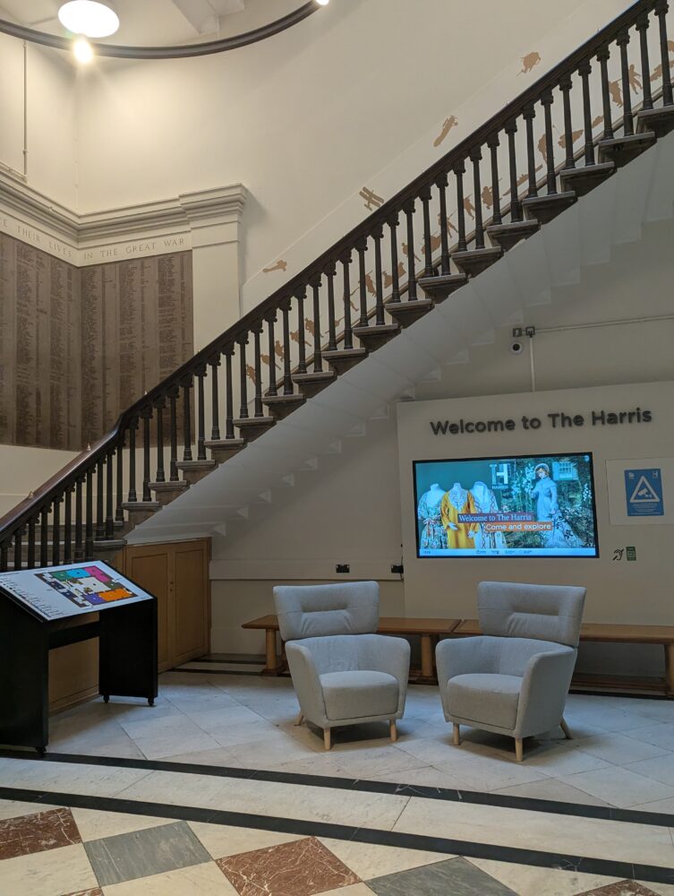 Modern lobby with two grey armchairs under a staircase. A digital sign reads "Welcome to The Harris," next to a wall-mounted display screen.