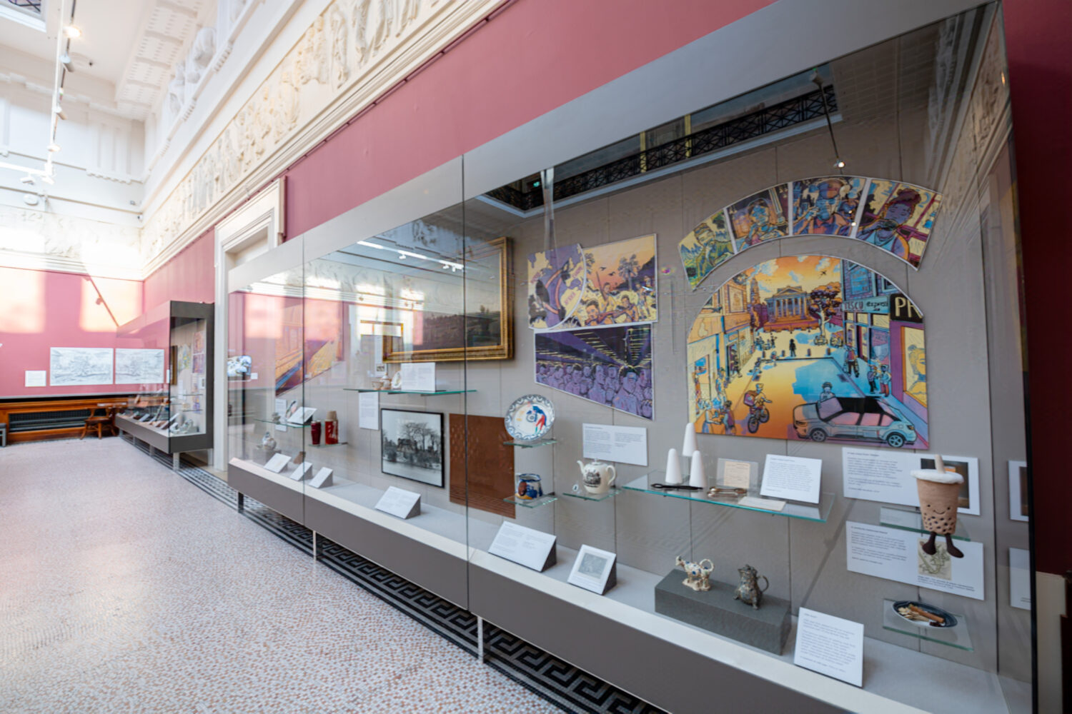 Museum exhibit with colorful cityscape artwork, ceramics, and historical photos. The bright room has a high ceiling and intricate molding, conveying a sense of history and culture.