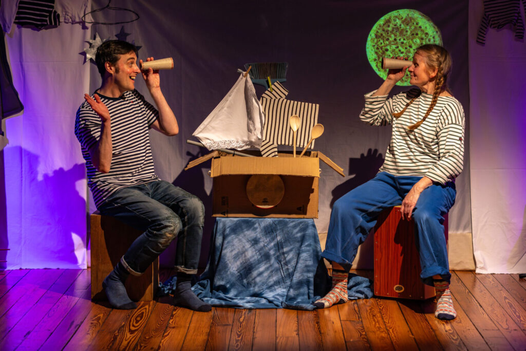 Image of two performers sat on boxes, both holding a telescope and looking at each other.