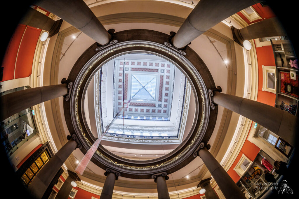 The image shows a grand, ornate museum interior with a high ceiling and skylight. Artwork adorns the red walls, and a decorative railing encloses a central exhibit.