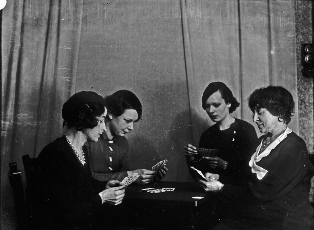 A black and white photograph of four people playing cards around a black circular table.