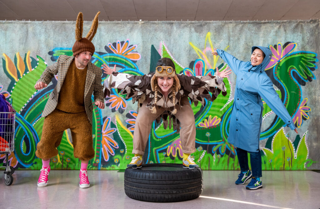 Three performers dressed as animals stood in front of a colourful backdrop and stood on a tire.