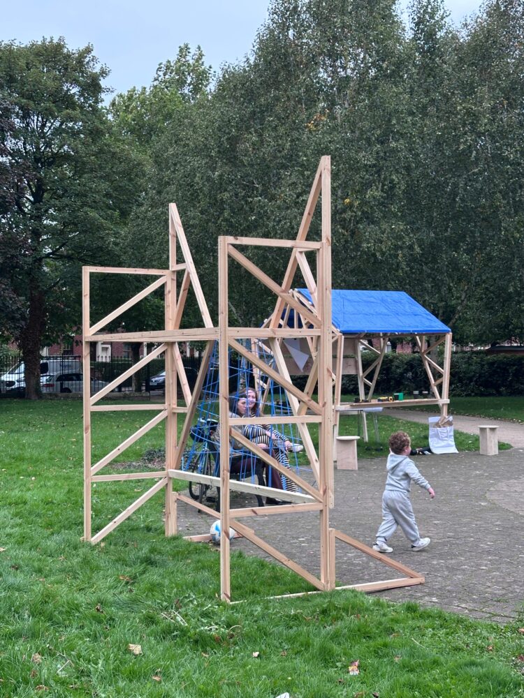 Image of a wooden climbing frame structure with a few people interacting with it in a park.