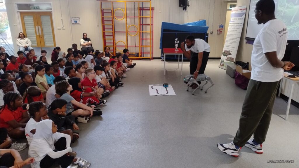 Image of a presentation in a school hall to school children with two instructors holding a robot dog.