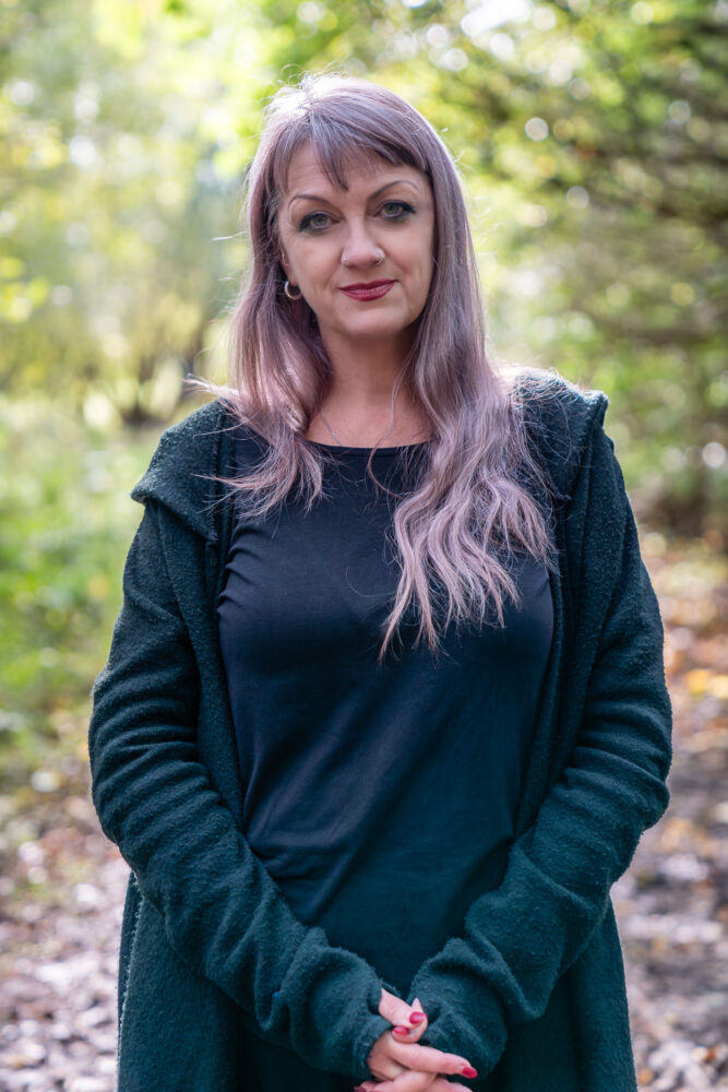 Image of a person stood in the forrest with purple hair and wearing a dark green long sleeved top.