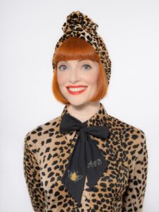 Studio portrait of a person with a bright red bob hairstyle, wearing a leopard-print outfit with a matching headwrap and a black bow at the neck against a plain light background.