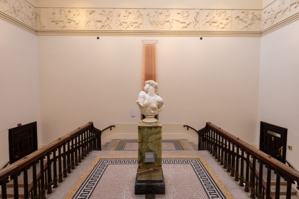 A marble bust atop a patterned pedestal stands at the center of a grand staircase in an art gallery. Sculptures and abstract wall art are displayed around.