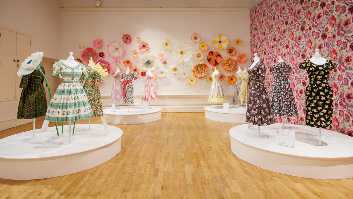 Image of an exhibition gallery filled with colourful dresses and flowers.