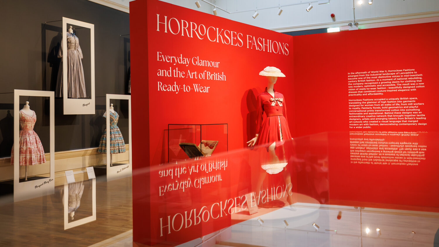 Image of an exhibition gallery filled with colourful dresses including a red one on a mannequin wearing a stylish white hat.